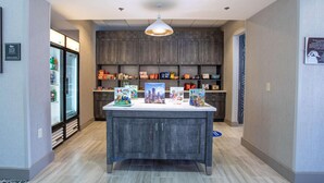 A hotel lobby with a central island displaying children's books, a refrigerator stocked with beverages, and a modern pendant light. - Homewood Suites by Hilton Leesburg (Leesburg)