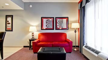 A hotel room with a red sofa, two framed artworks on the wall, a black ottoman, a standing lamp, and a window with curtains.