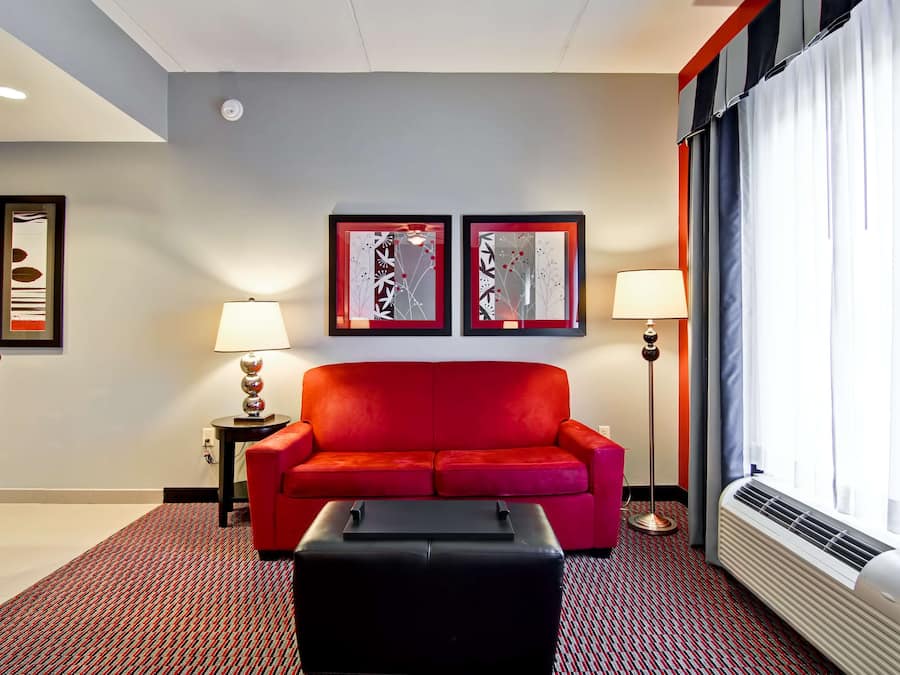A hotel room with a red sofa, two framed artworks on the wall, a black ottoman, a standing lamp, and a window with curtains.