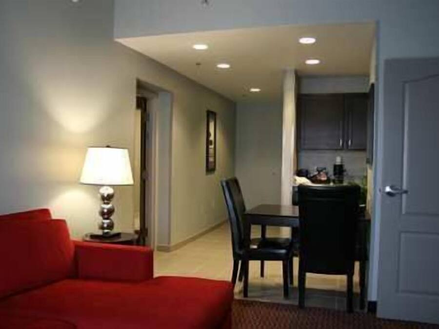 A compact living space with a red sofa, a dining table with chairs, a lamp, and a kitchen area visible through an open door.