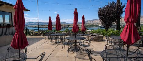An outdoor patio with round tables and chairs, red umbrellas, and a scenic view of a lake and mountains.