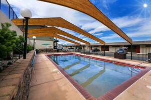 Una piscina con cubierta de madera y sillas de descanso, rodeada por un edificio con valla metálica.
