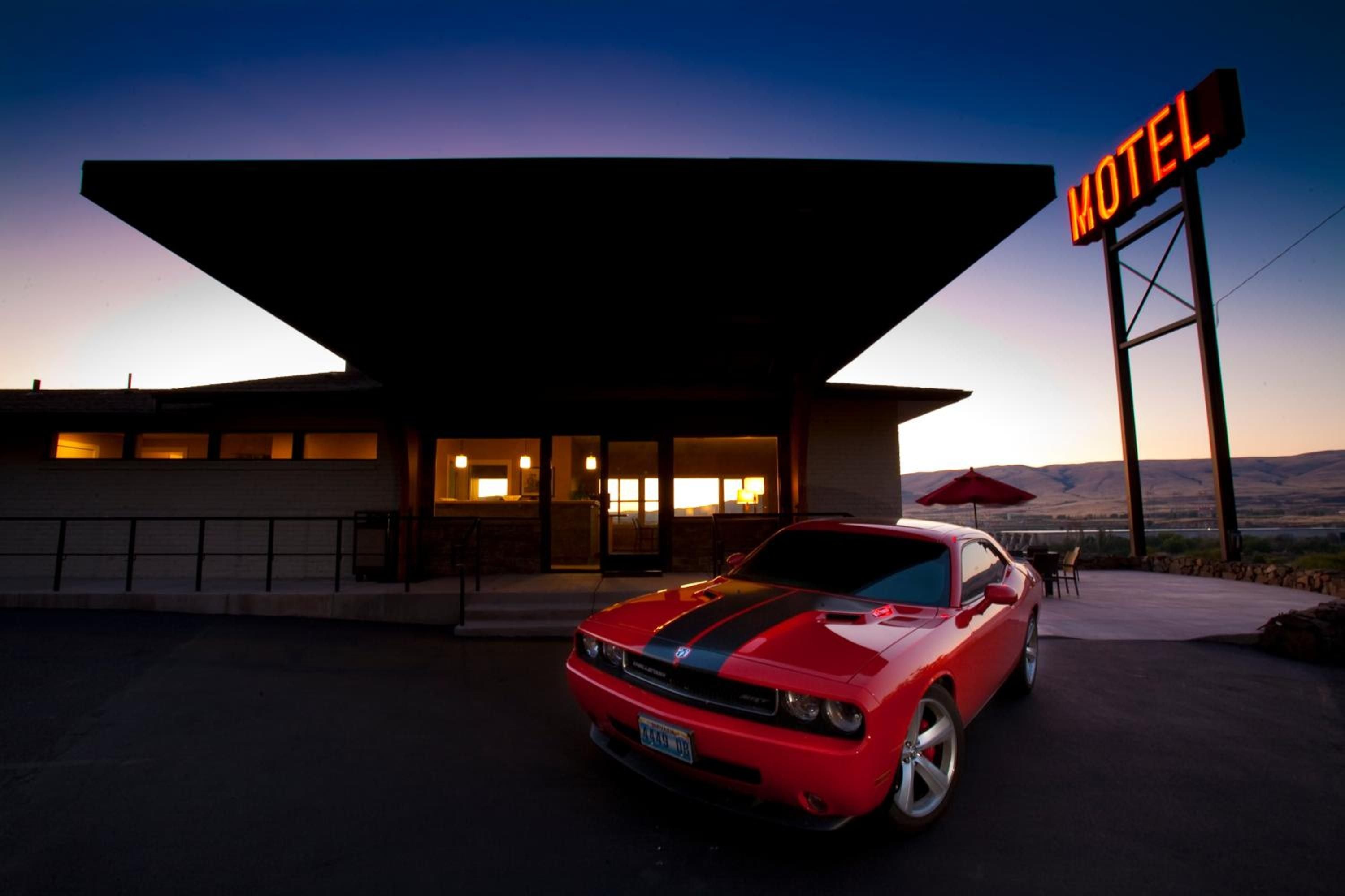 A red sports car parked in front of a motel with a large awning and a neon sign.