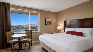 A hotel room with a large bed, a desk with two chairs, a window with a view of a bridge and hills, and a framed picture on the wall. - Celilo Inn (The Dalles)