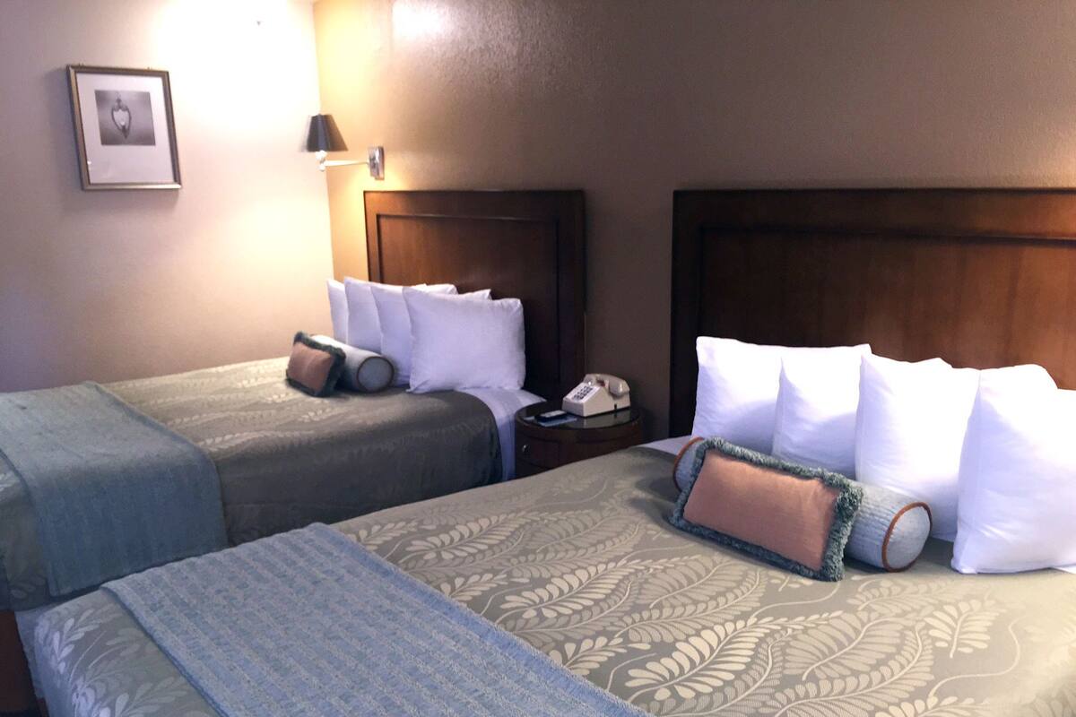 A hotel room with two beds, each with a wooden headboard, white bedding, and a bedside table with a lamp.