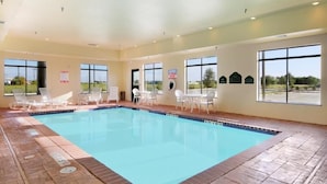 Piscina com espreguiçadeiras e mesas, janelas amplas e vista para o exterior.
