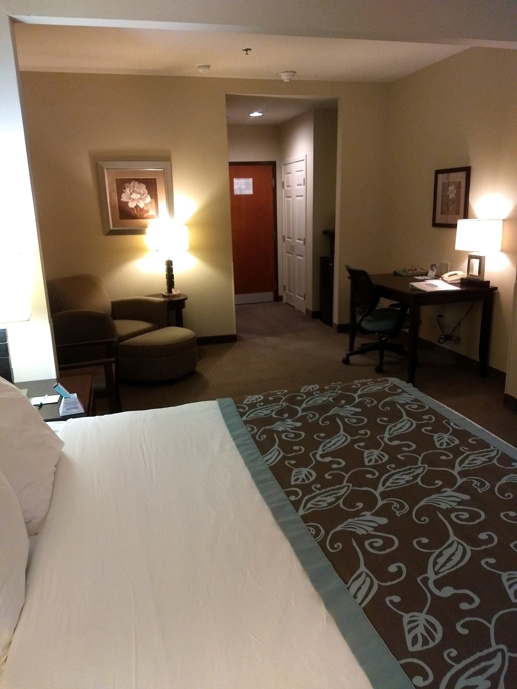 A hotel room with a bed, a desk, two chairs, a lamp, and a door leading to another room.
