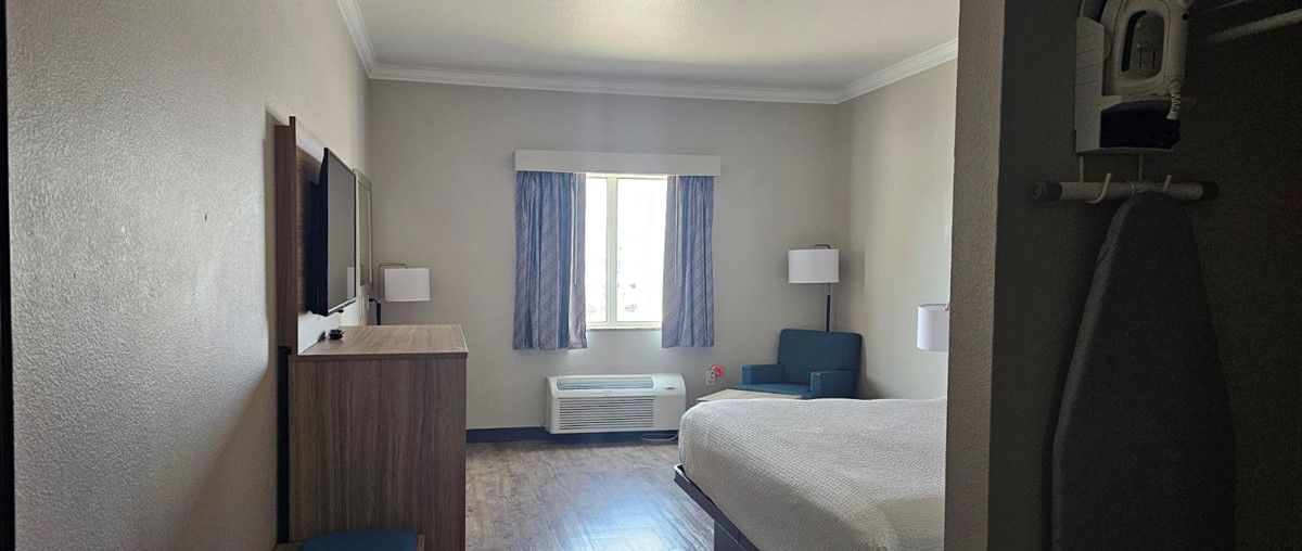 A hotel room with a bed, a desk, a chair, a TV, a window with curtains, and an air conditioning unit.