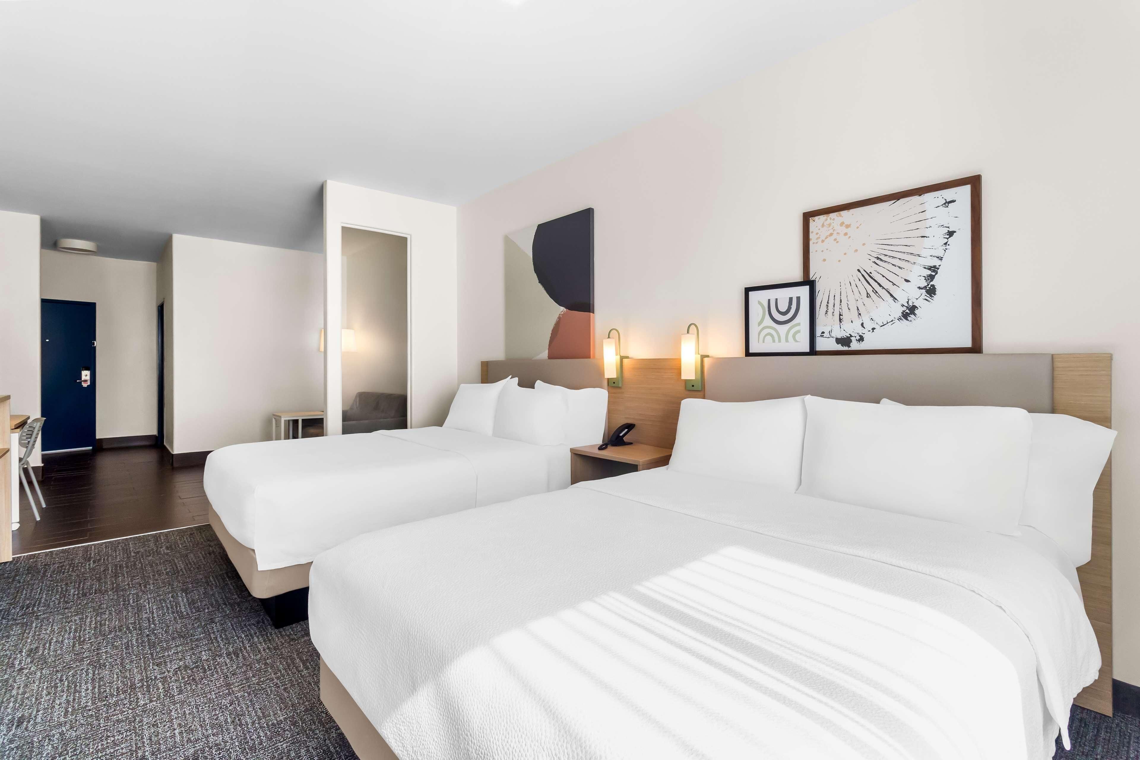 Une chambre d’hôtel avec deux lits, un bureau, une chaise, une petite table et un mur décoré d’œuvres d’art abstraites.