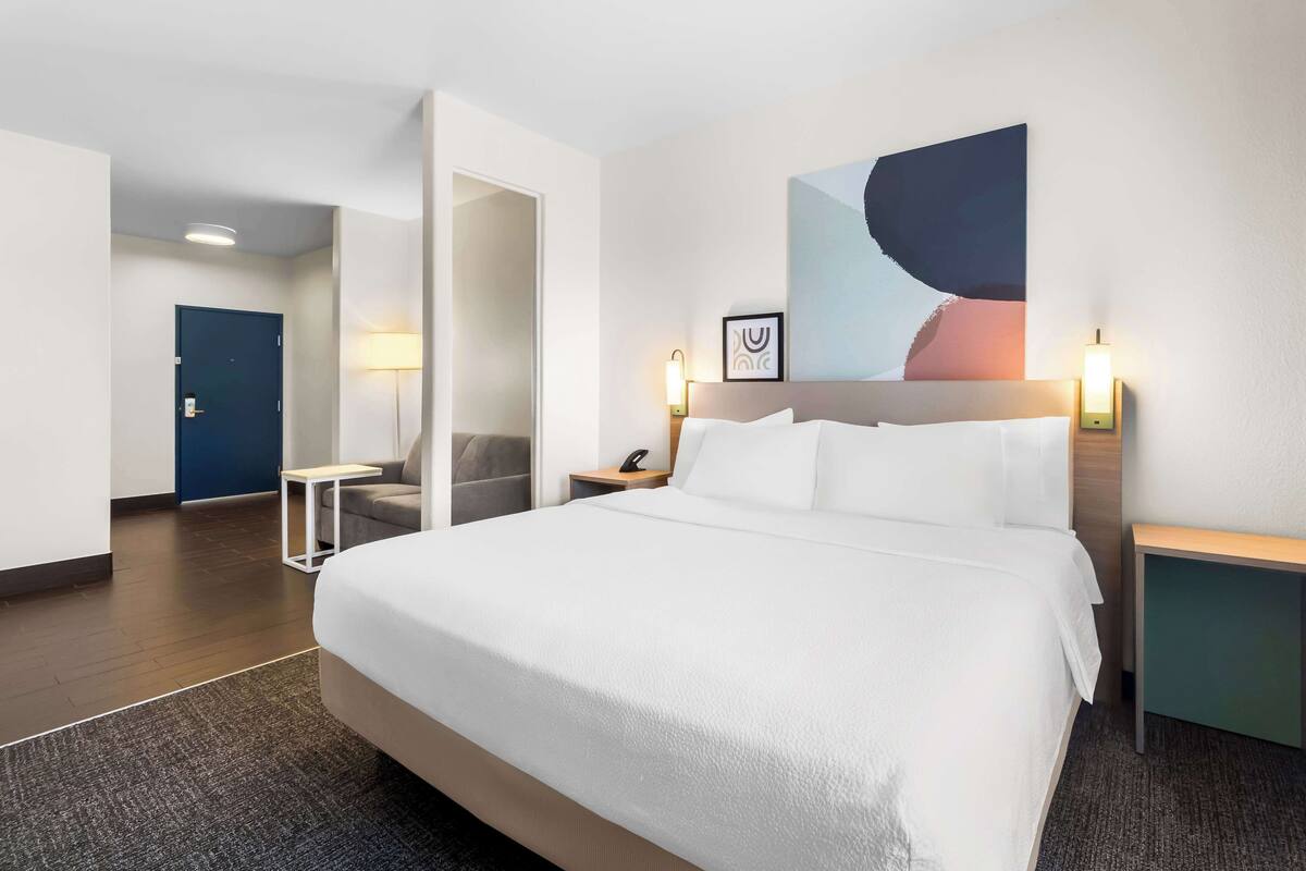 A hotel room with a large bed, a desk, a chair, and a door leading to another room.