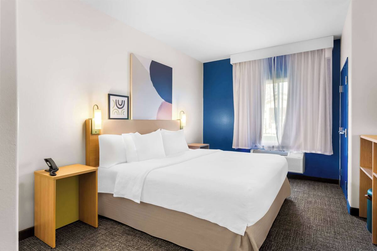 A hotel room with a bed, bedside tables, a wall-mounted artwork, a window with curtains, and a blue accent wall.
