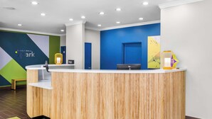 A reception area with a wooden reception desk, a bench, a wall with a geometric design, and a door. - Spark by Hilton Pearland (Pearland)