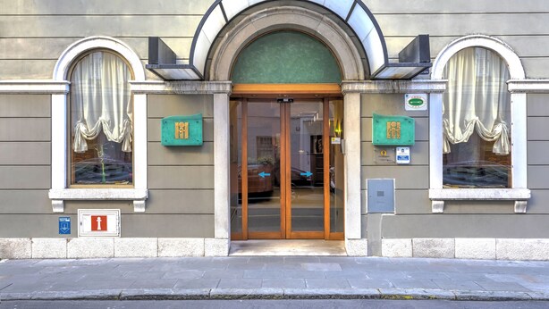 A hotel entrance with a glass door, two green service boxes, and a fire alarm sign. - Hotel Italia (Trieste)
