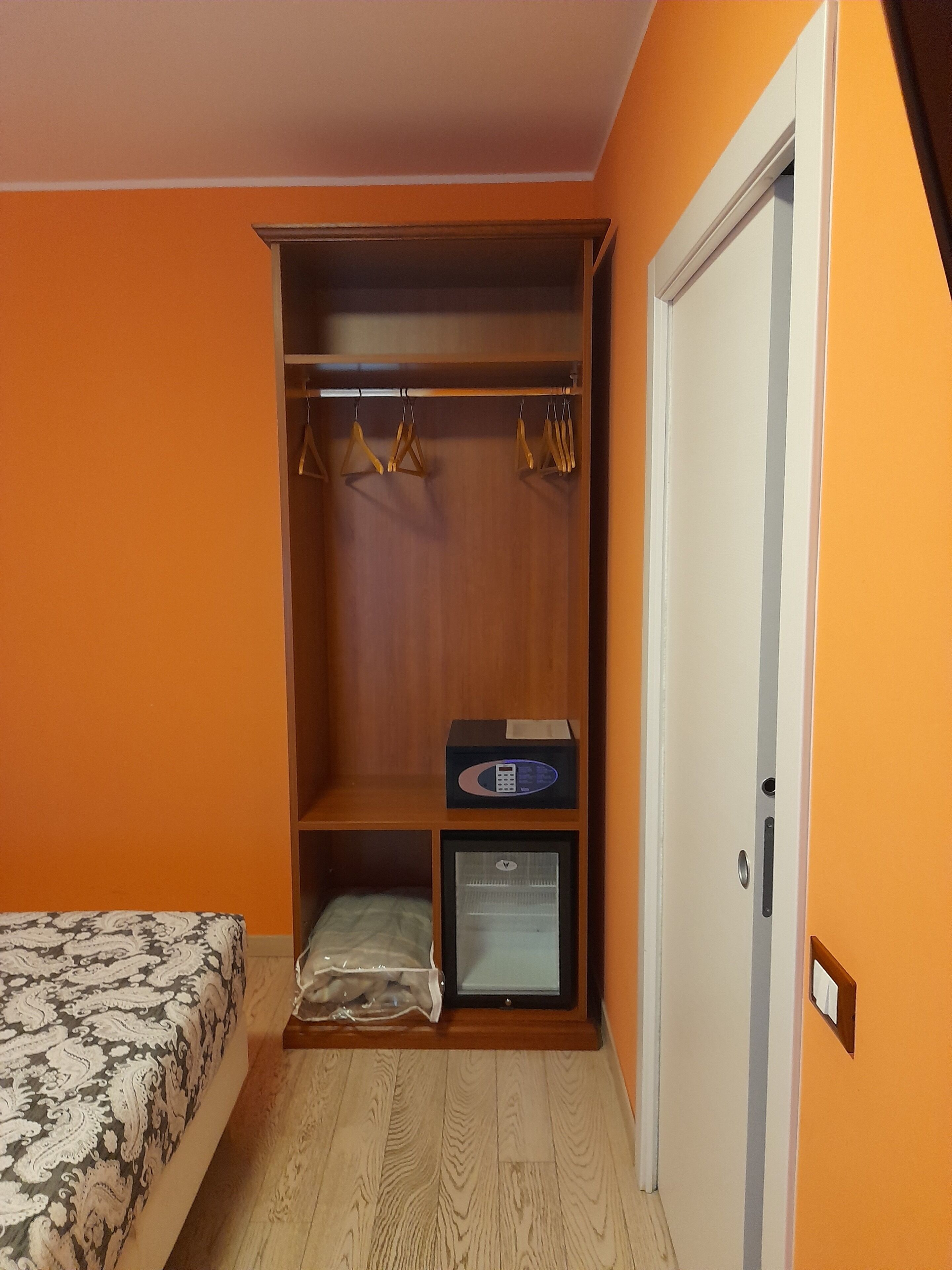 Un dormitorio con una pared naranja, un armario de madera, una cama con ropa de cama estampada y un refrigerador pequeño.