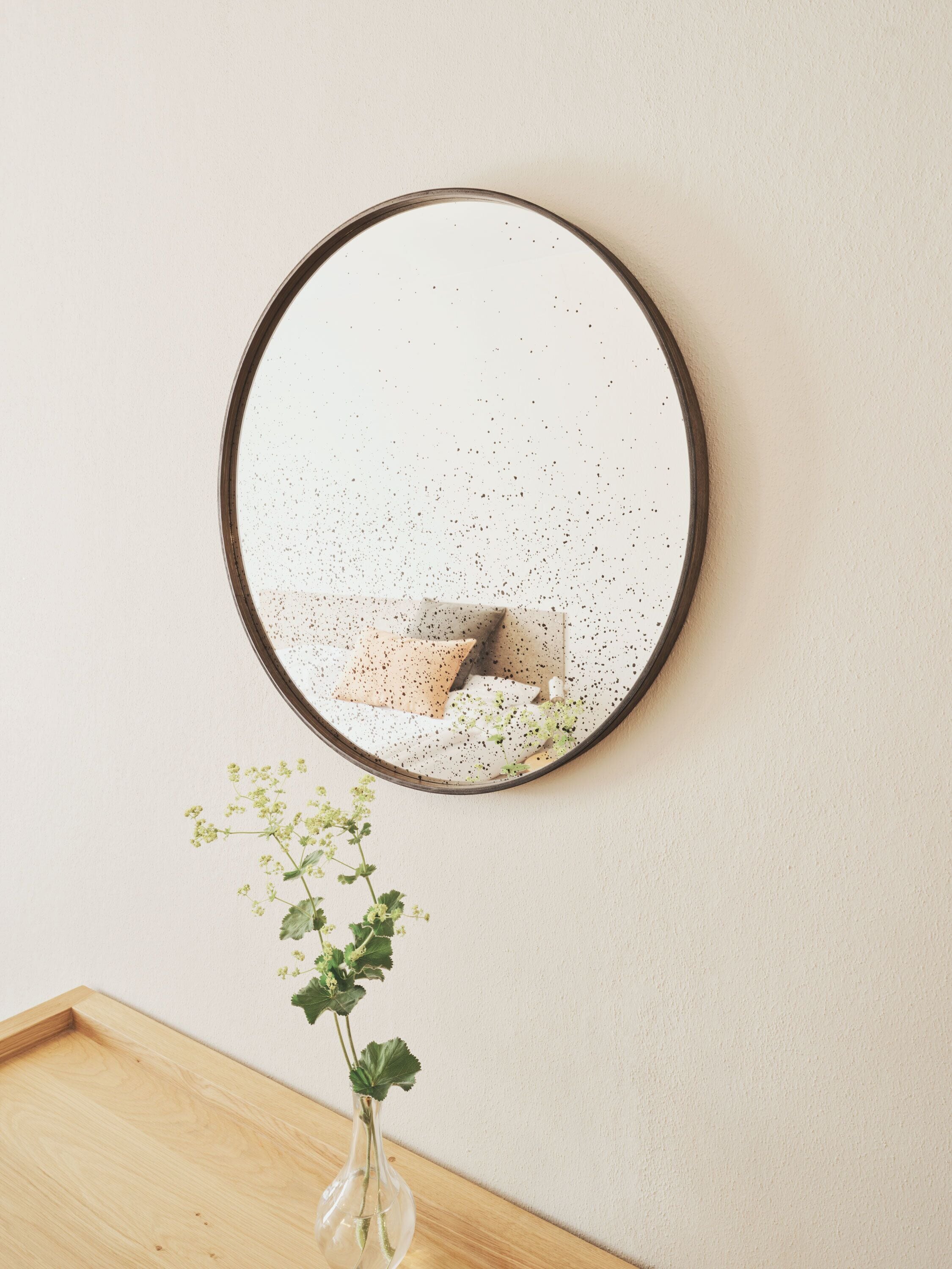 A round mirror with a speckled surface mounted on a wall. Below the mirror, there is a wooden surface with a clear glass vase containing green foliage.