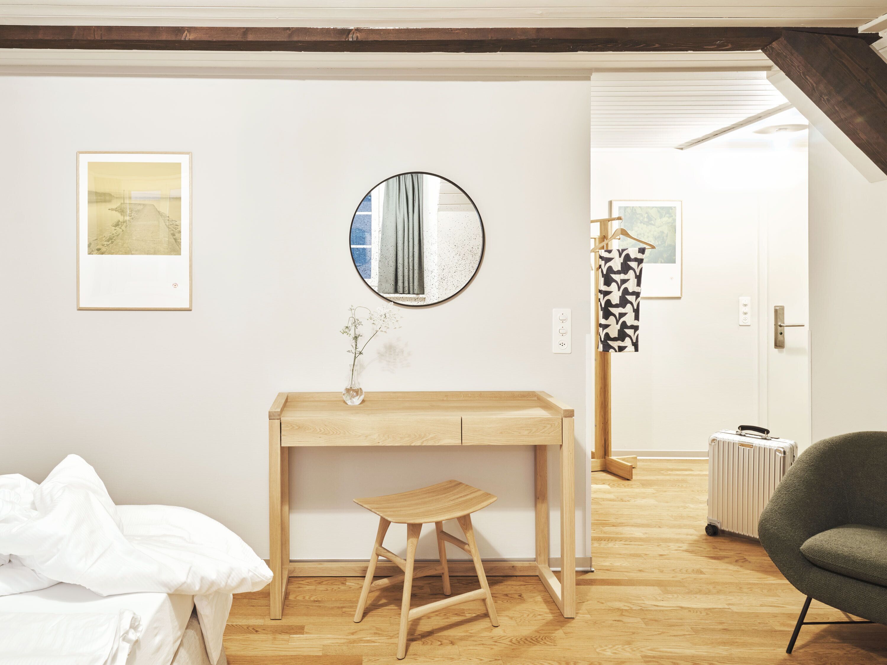 Una habitación con un escritorio de madera, un espejo redondo, un taburete pequeño, una cama con ropa blanca y un radiador.