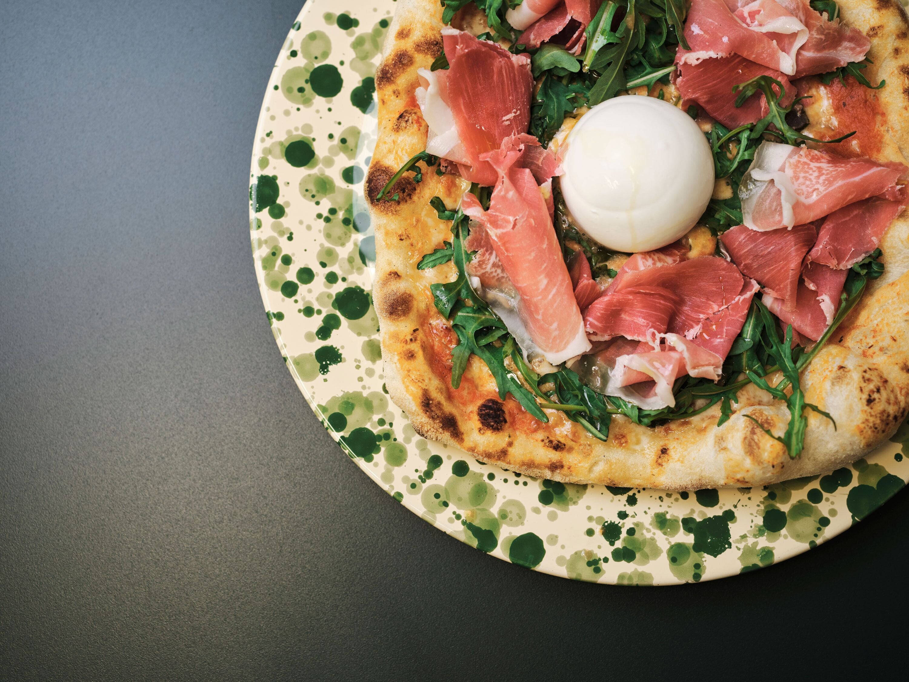 A pizza with prosciutto and arugula on a speckled plate.