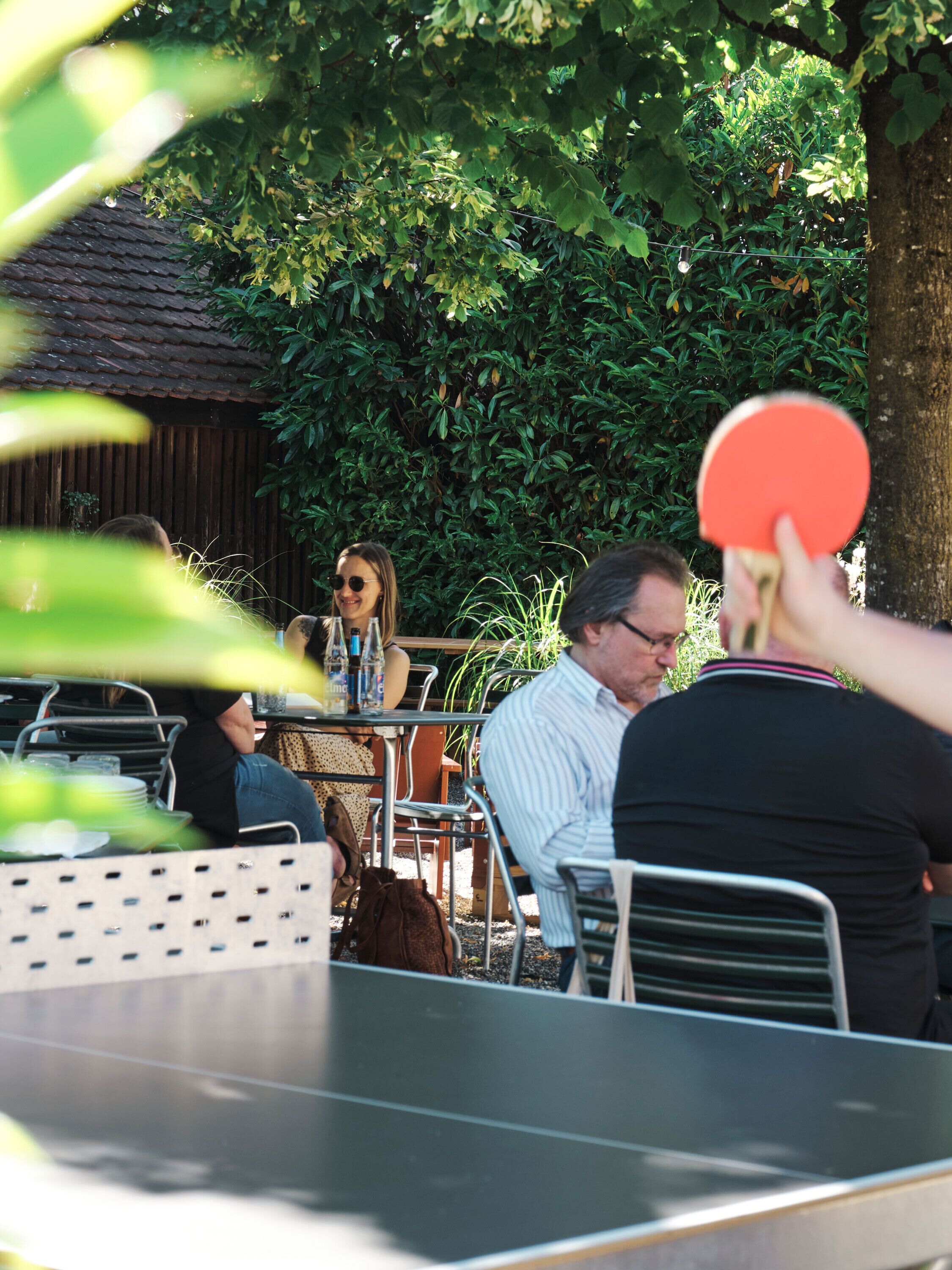 An outdoor dining area with metal chairs and tables, surrounded by greenery and a wooden fence.