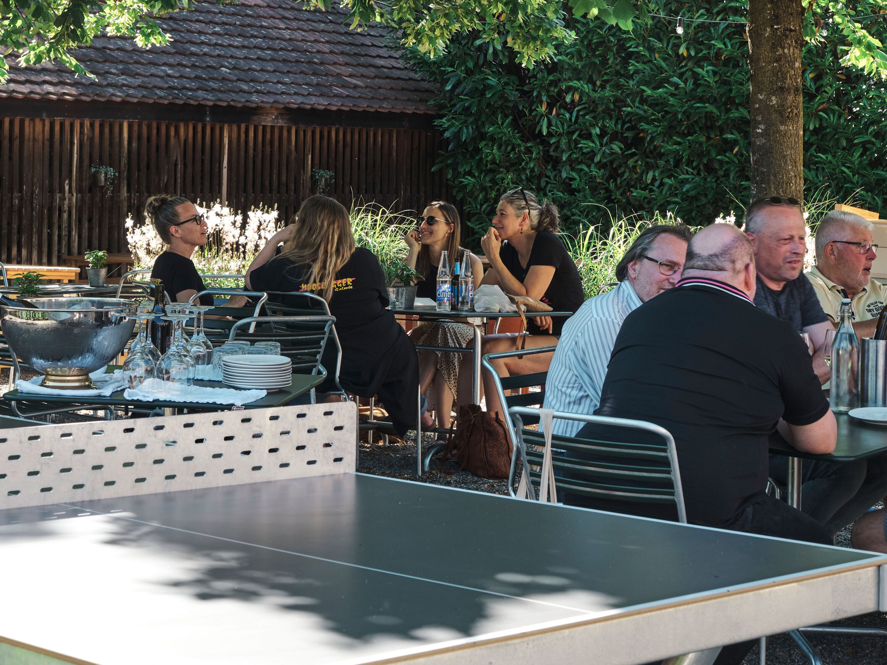 Un área de comedor al aire libre con mesas y sillas de metal, rodeada de follaje y una cerca de madera.