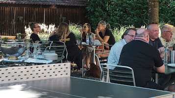 Un área de comedor al aire libre con mesas y sillas de metal, rodeada de follaje y una cerca de madera.