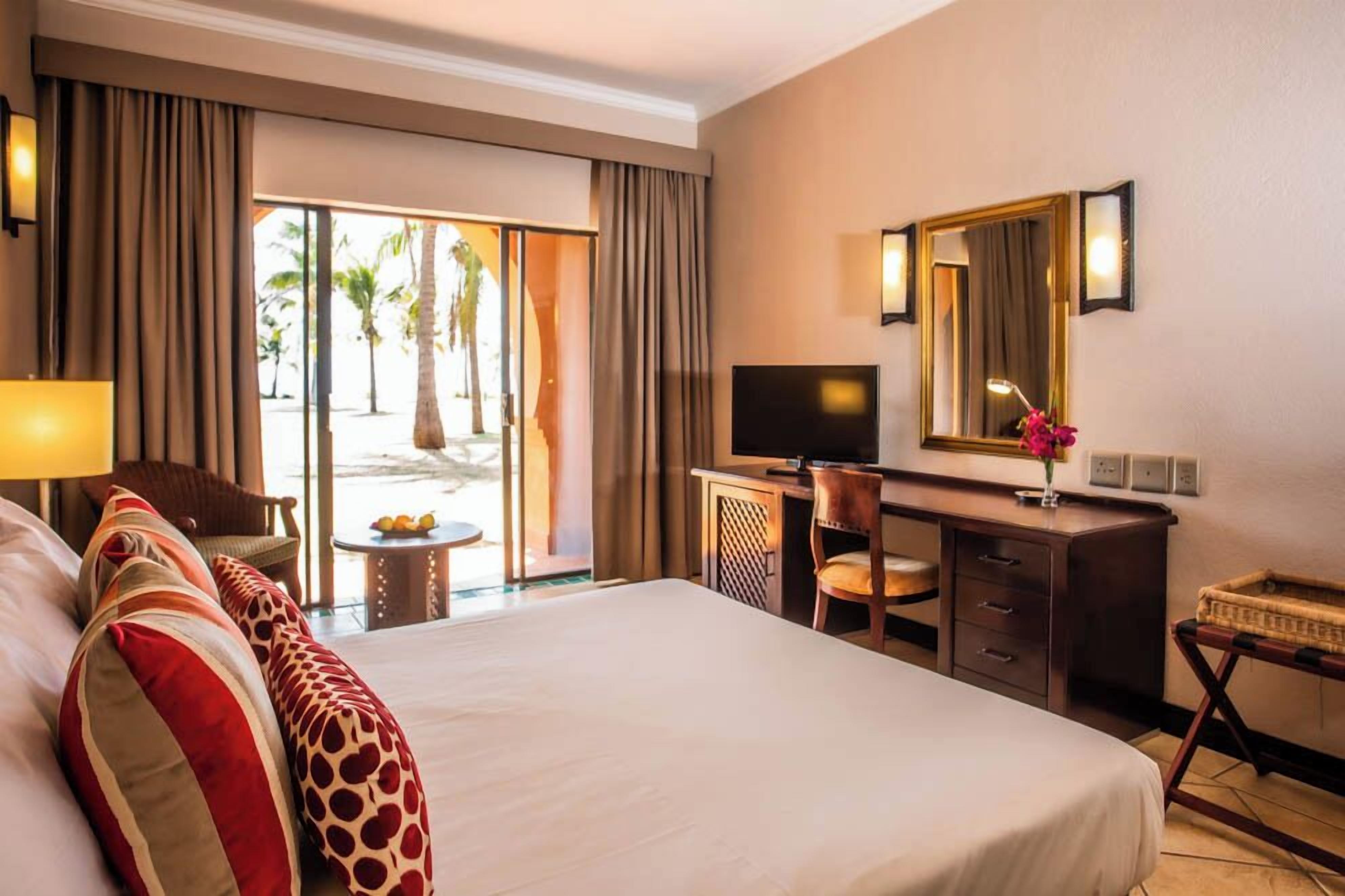 A hotel room with a large bed, a desk with a TV, a chair, and a view of palm trees through the window.