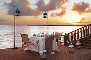 A sunset view from a wooden deck with a table set for two, two chairs, and two lanterns. - Avani Pemba Beach Hotel (Pemba)