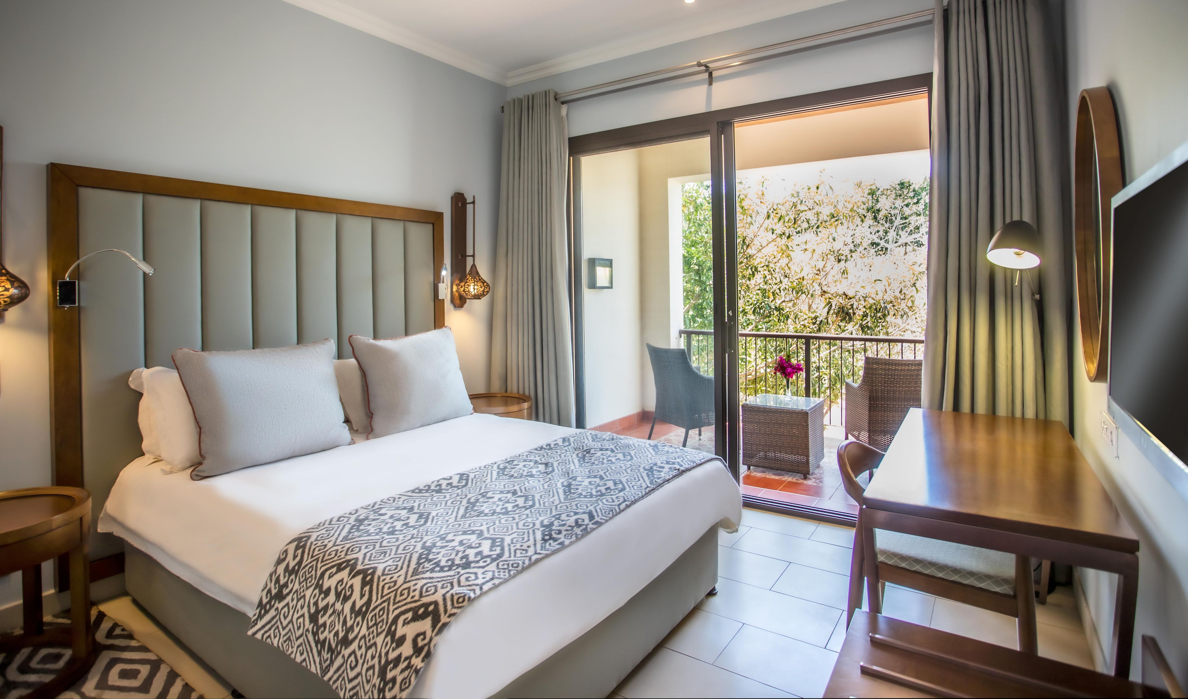 Habitación de hotel con cama, escritorio, silla, televisión y balcón con vista a la vegetación.