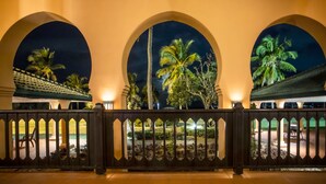 Vue nocturne d’un hôtel avec trois fenêtres en arc donnant sur un jardin planté de palmiers.