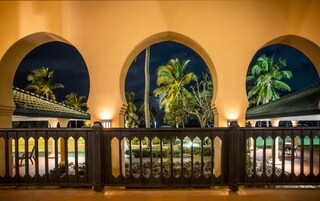 Vista nocturna de un hotel con tres ventanas arqueadas que dan a un jardín con palmeras.