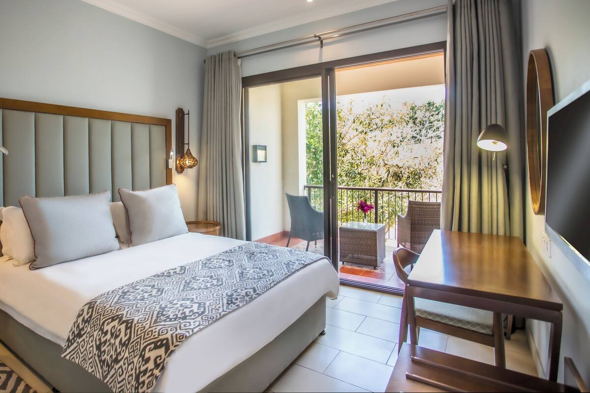 Habitación de hotel con cama, escritorio, silla, televisor y balcón con vista a árboles.