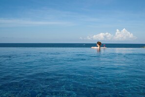 Una persona relajándose en una piscina infinita azul cristalina con vista al océano y un barco a lo lejos.