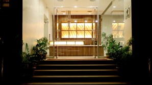 Vestíbulo de hotel con mostrador de recepción, una escalera que sube a los pisos superiores y plantas en macetas.
