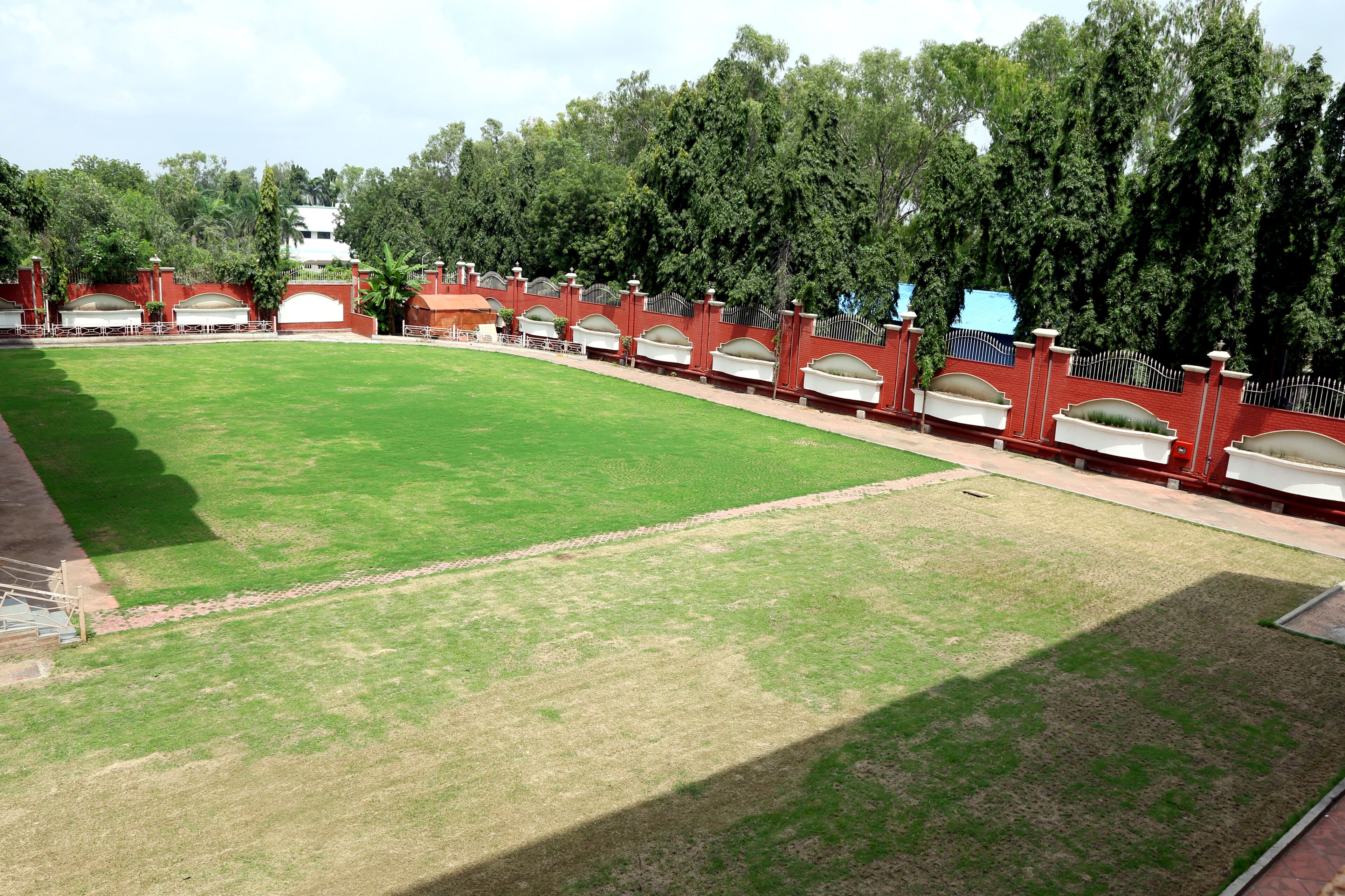 A large, open grassy area with a curved red brick wall and white arches, surrounded by trees and a white building in the background.