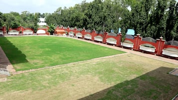 A large, open grassy area with a curved red brick wall and white arches, surrounded by trees and a white building in the background.