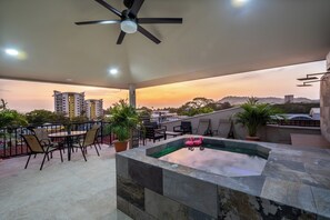 Una terraza en la azotea con jacuzzi, zona de estar y vistas a edificios de la ciudad al atardecer.
