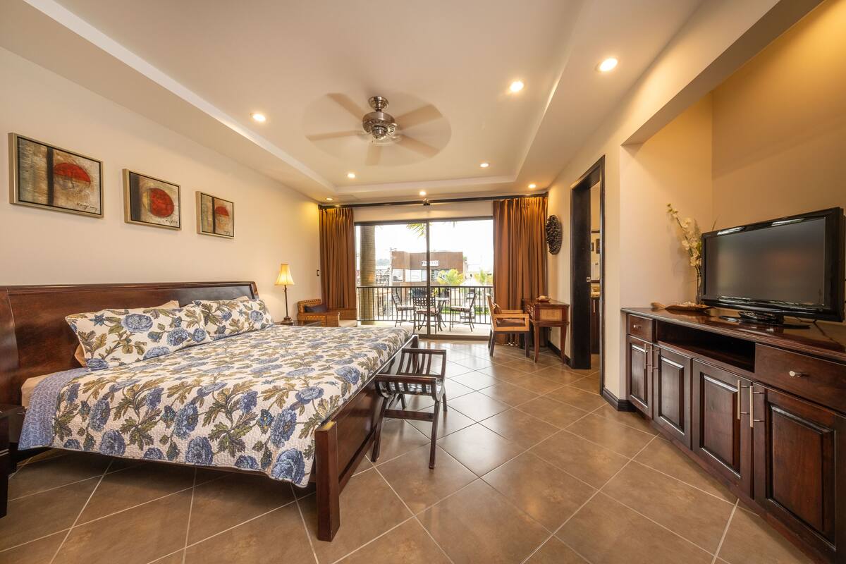 A hotel room with a bed, a TV, a balcony with a table and chairs, and a ceiling fan.