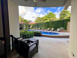 A pool area with lounge chairs and a table, surrounded by greenery and a clear sky.