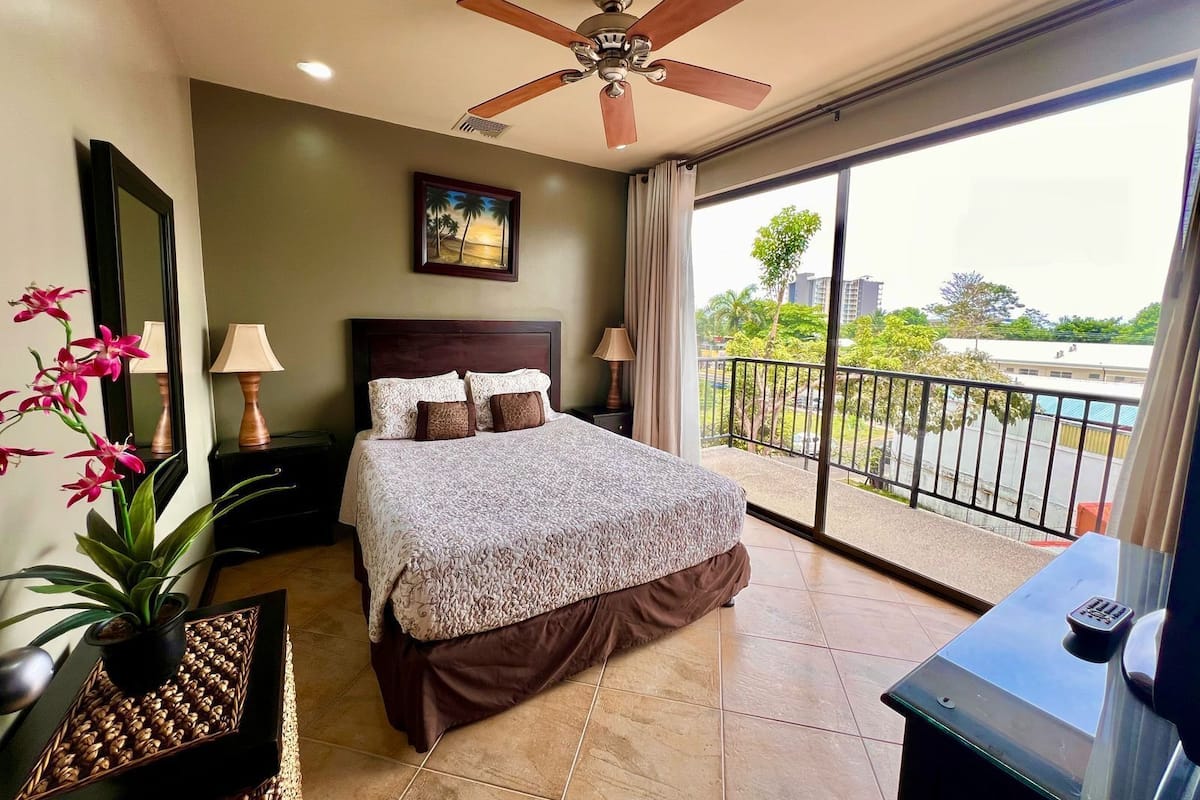 A hotel room with a large bed, a ceiling fan, a balcony with a view, and a painting on the wall.