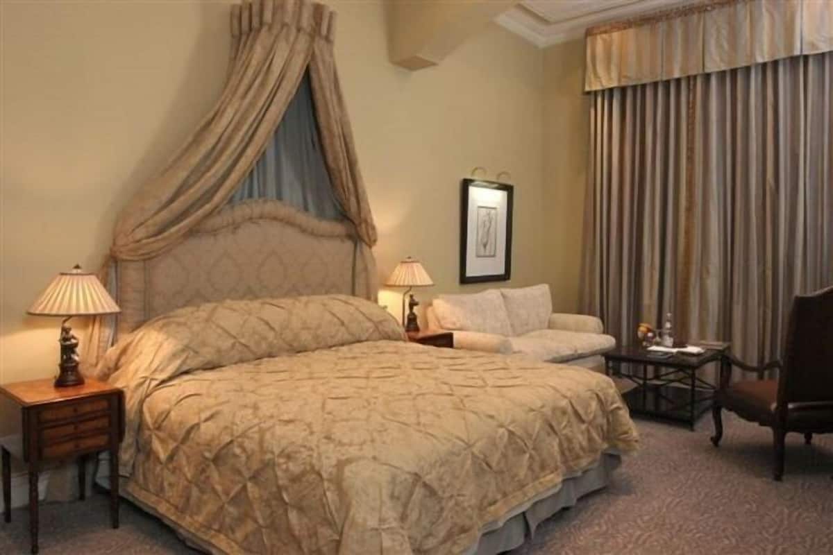 Ein Schlafzimmer mit einem großen Bett, einem Nachttisch mit Lampe, einem Sessel, einem gerahmten Bild an der Wand und einem Fenster mit Vorhängen.
