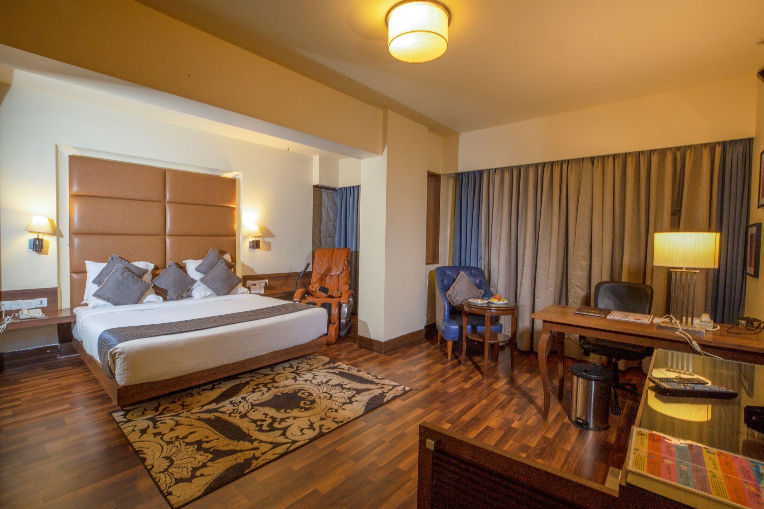 A hotel room with a large bed, a desk, a chair, a small table, and a lamp.