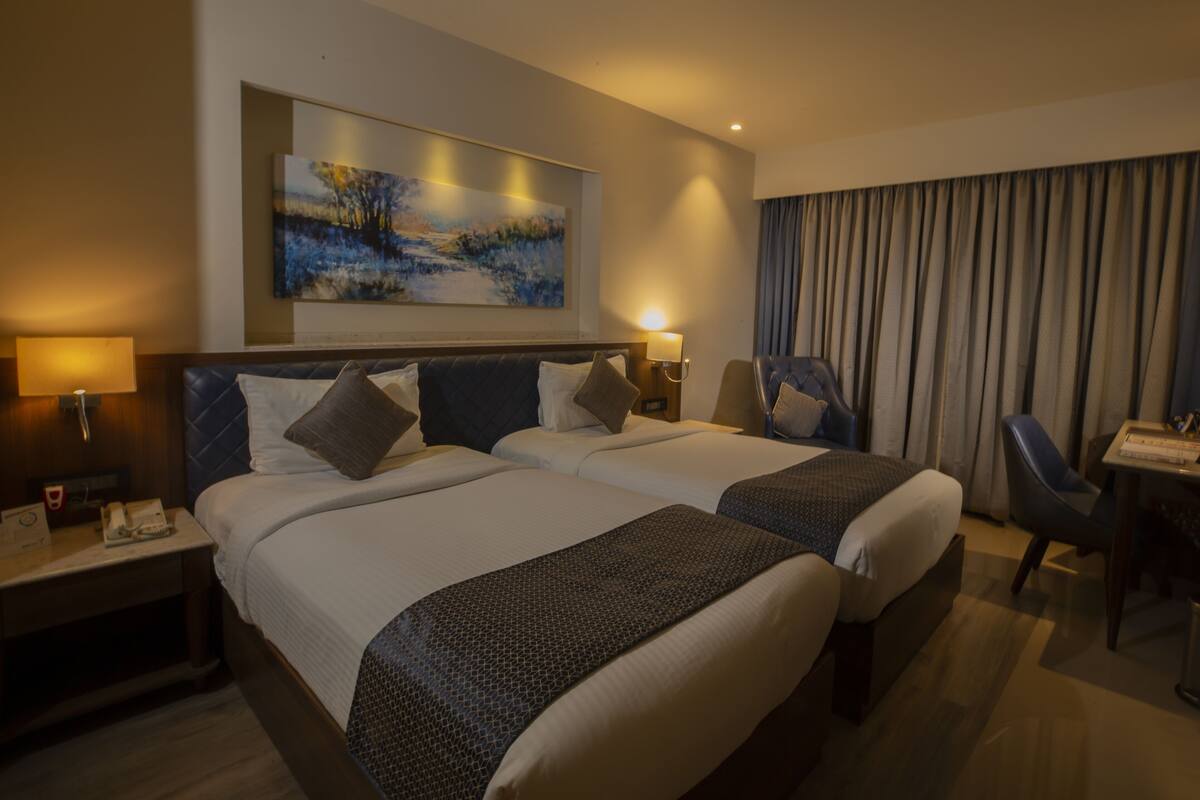 A hotel room with two beds, a desk, a chair, a painting on the wall, and a window with curtains.