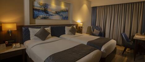 A hotel room with two beds, a desk, a chair, a painting on the wall, and a window with curtains.
