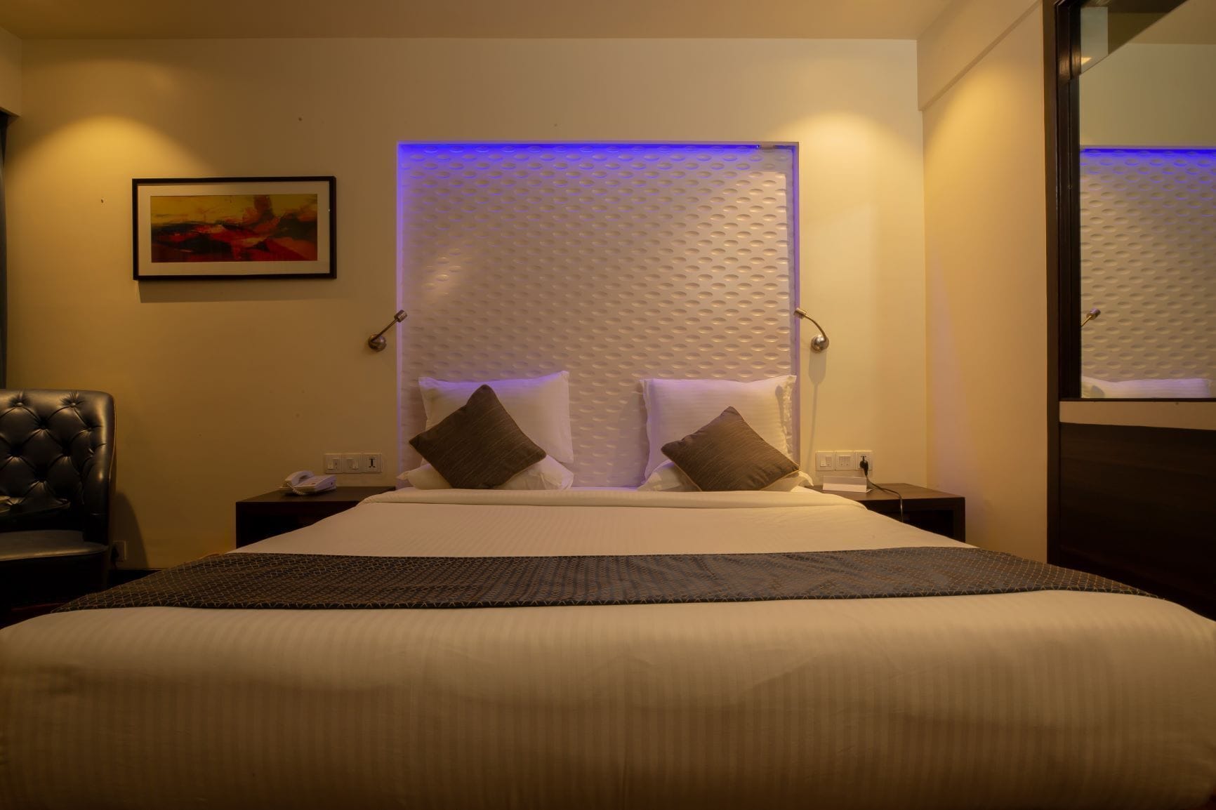 A hotel room with a large bed, two bedside tables, a chair, a framed picture on the wall, and a textured headboard.