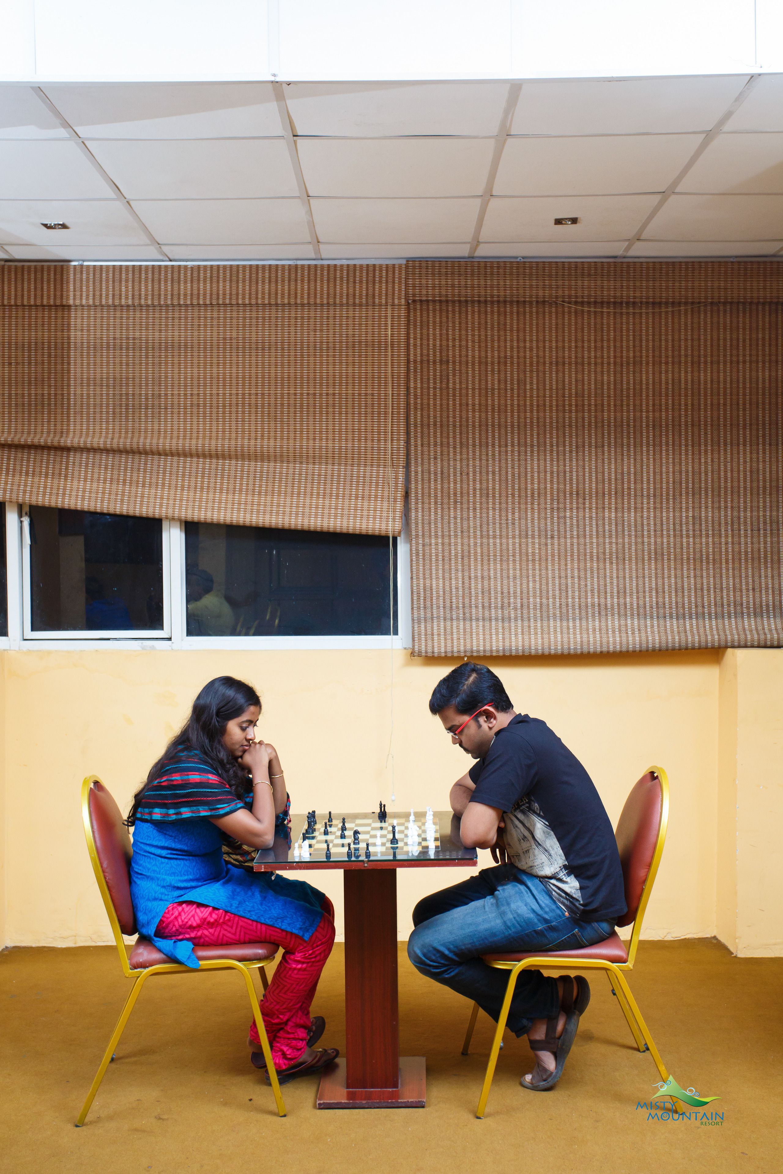 Two people playing chess at a table in a room with yellow walls and a window with blinds.