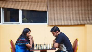 Two people playing chess at a table in a room with yellow walls and a window with blinds.