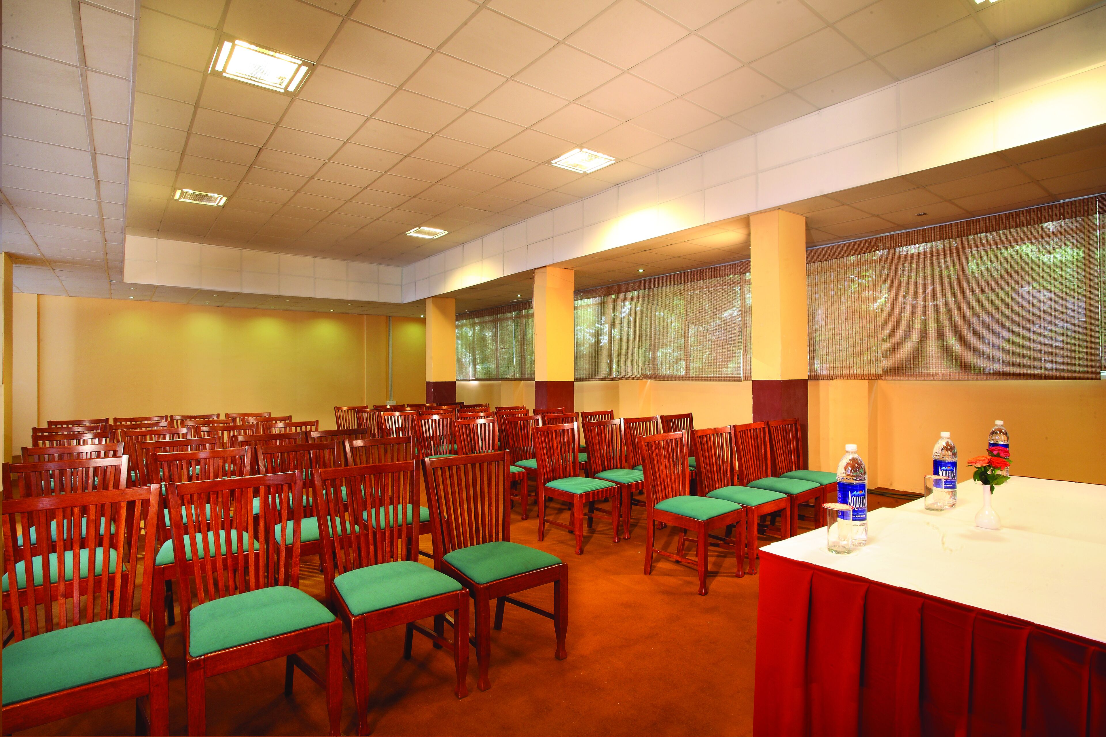 A conference room with rows of wooden chairs, a white table with water bottles, and a view through large windows.