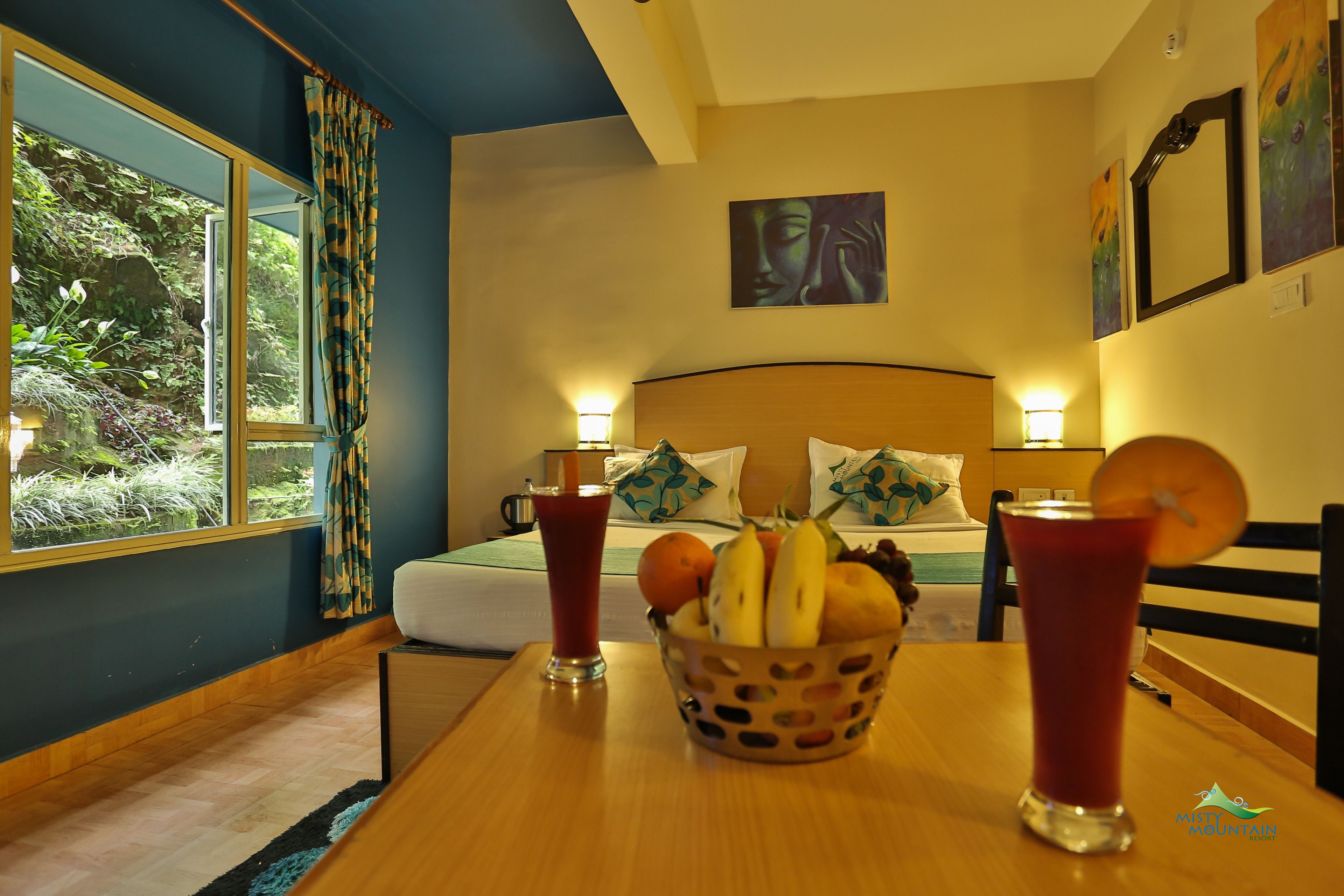 A hotel room with a bed, a table with fruit and drinks, and a view of greenery outside.
