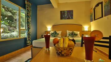 A hotel room with a bed, a table with fruit and drinks, and a view of greenery outside.