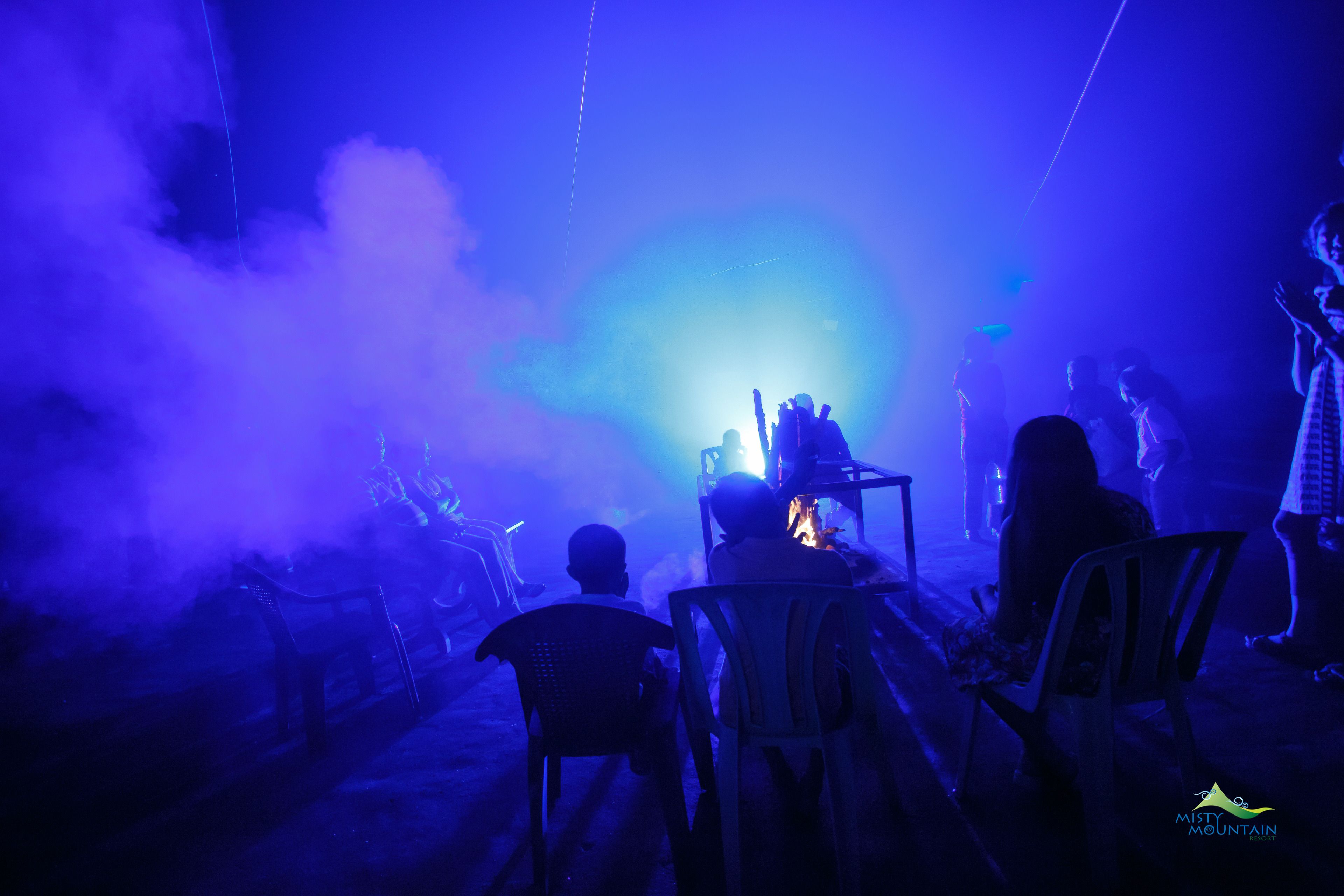 A dimly lit room with chairs arranged around a central table, a visible fire, and a smoke effect.