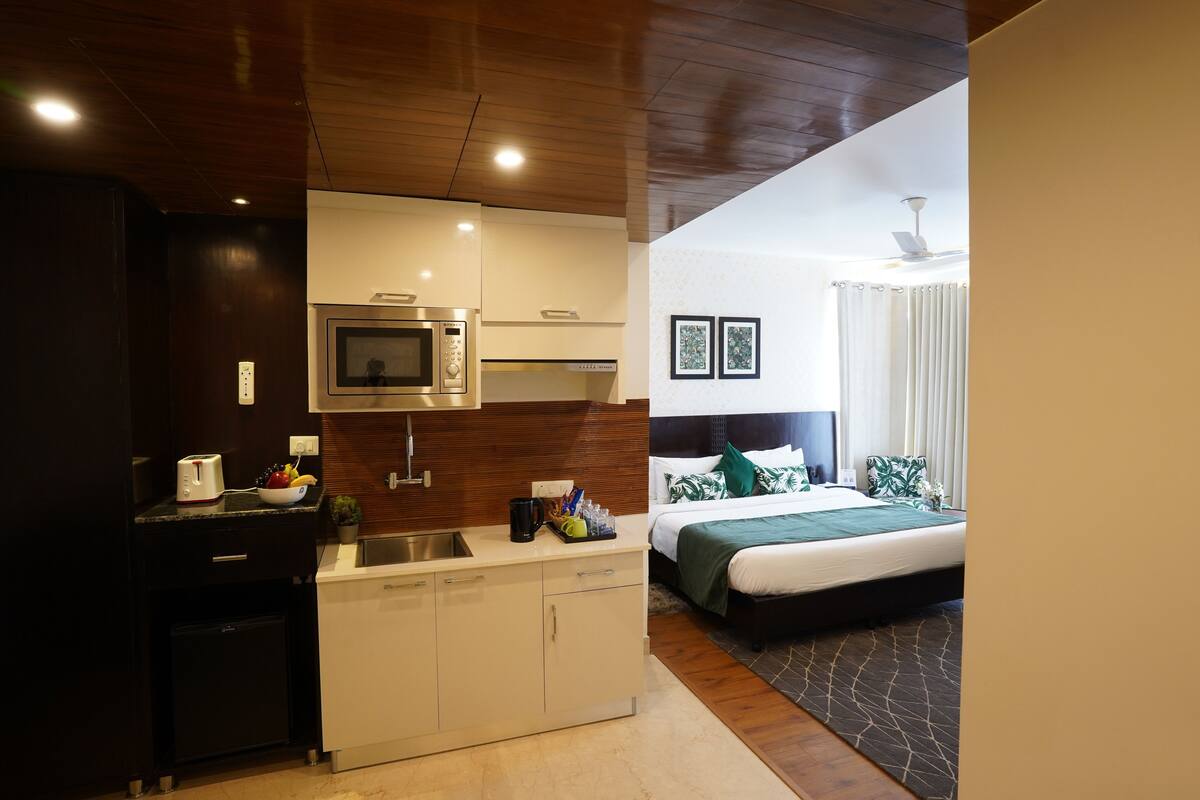 A compact hotel room with a kitchenette, a bed with green bedding, and a ceiling fan.